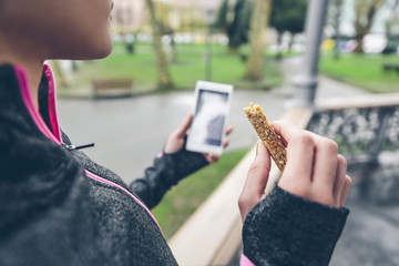 Woman eating cereal bar after training and holding smart phone