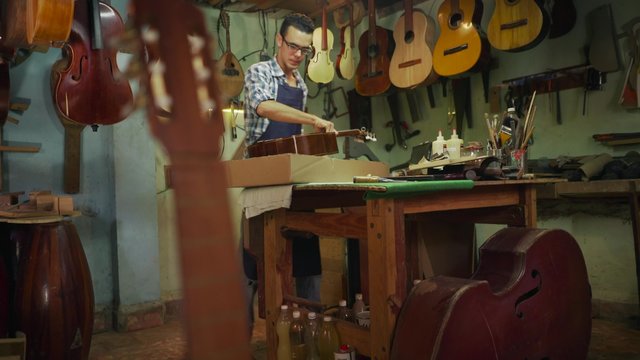 Lute maker shop and acoustic music instruments: a young adult artisan fixes an old classic guitar, then stores it in a cardboard case for his client. Crane shot, Wide shot