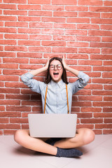 Beautiful young dark-haired girl in casual clothes with laptop, looking in camera and showing surprise, sitting cross-legged against brick wall