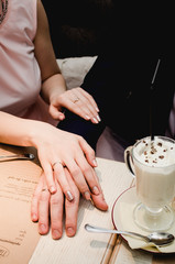 Loving woman hold man hand in her hands. Just married couple showing up wedding rings. Near Cup of latte coffee with chocolate on saucer with spoon stand on on brown wooden table in cafe