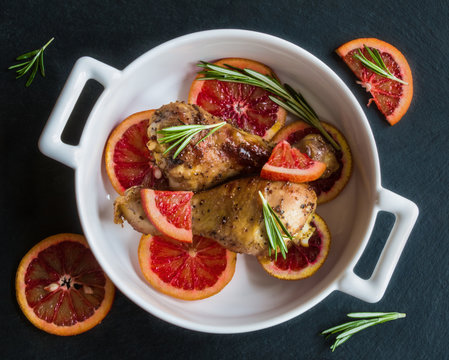 Roasted chicken legs on slices of red oranges in white baking dish. Black slate background. Cooked with the sauce: orange juice, mustard, olive oil and honey. 