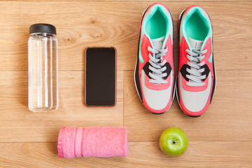 red and grey sneakers with grey shoelaces and red towel, green apple, bottle with water, mobile phone with white headphones on wooden background indoors