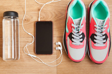 red and grey sneakers with grey shoelaces and bottle with water, mobile phone with white headphones on wooden background indoors
