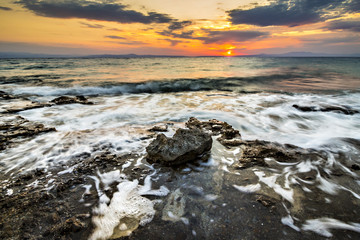 Rising sun, stone and the sea with wave on sunset time at the coast of Aegean seashore in Greece