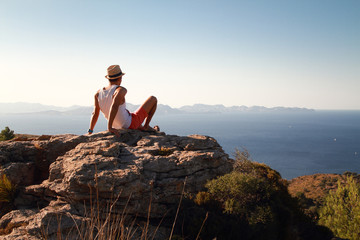 Naklejka na ściany i meble Contemplative young man sitting on the rocks on the top of the mountain at sunset. Looking at distance with the blue sea in front. Wearing a straw hat in summer.