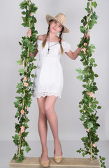 full-length Beautiful young leggy blonde in a little white dress and white cowboy hat on a swing, wooden swing suspended from a rope hemp, rope wrapped vine and ivy