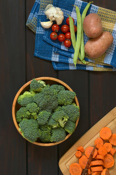 Fresh raw broccoli florets in wooden bowl with carrot slices on wooden board, potato, green bean, cherry tomato and garlic on kitchen towel, photographed on dark wood with natural light