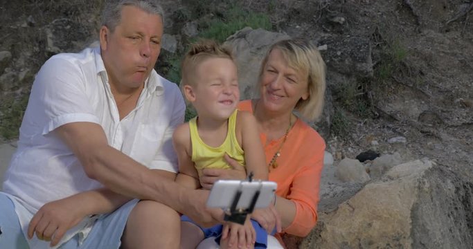 Grandson and grandparents having fun while taking outdoor cellphone selfie using monopod. They making funny faces and laughing