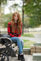 Fototapeta na wymiar Young grumpy redhead woman in red plaid jacket and blue jeans sitting on park bench with blurred park background