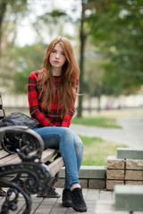 Fototapeta na wymiar Young curious redhead woman in red plaid jacket and blue jeans sitting on park bench with blurred park background
