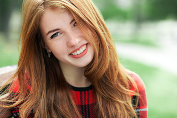 Closeup portrait of young laughing redhead woman in red plaid jacket with blurred park background