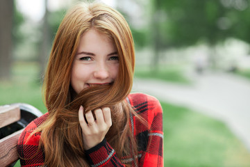 Closeup portrait of young smiling redhead woman in red plaid jacket touching her hair with blurred park background