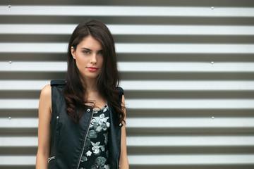 Closeup portrait of young beautiful brunette woman in black leather jacket posing outdoors against urban style background of metal strips