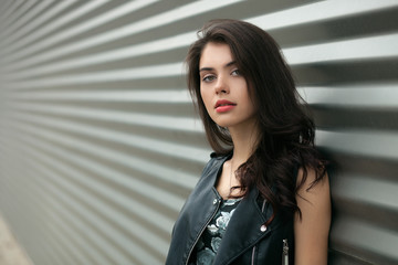 Closeup portrait of young beautiful brunette woman in black leather jacket posing outdoors against urban style background of metal strips