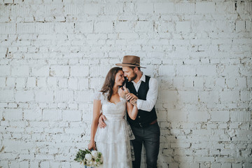 girl with a wreath of white flowers on the head in a white wedding dress and a bearded man in a suit and a hat are holding a bouquet of white flowers and green against a white brick wall
