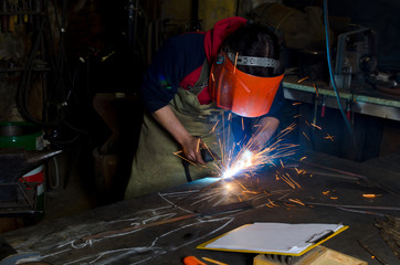 a man in a mask welds metal parts inside. On the table a notebook with a blank page