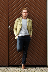 A fashionable man in his 20s wearing a yellow jacket and dark pants, leaning against a big wooden...