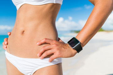 Fit bikini woman wearing wearable tech smartwatch. Closeup of slim abs stomach and beachwear on of...