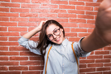 Beautiful young dark-haired girl in casual clothes and eyeglasses posing, smiling and making selfie, standing against brick wall