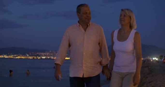 Steadicam shot of mature couple having romantic walk on the beach in the evening and taking selfie shot at smartphone.