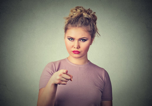 angry young woman pointing at camera isolated on grey wall background