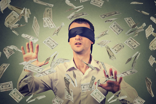 Blindfolded young businessman trying to catch dollar bills banknotes flying in air