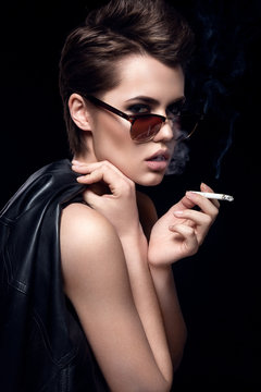 Fashion model smoking cigarette wearing sunglasses. Sexy woman portrait over dark background. Attractive fashion girl posing. Hairstyle, haircut
