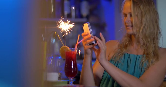 Young blond woman with smart phone taking picture of cocktail with sparkler and then making selfie with it. When fire going down she having a drink