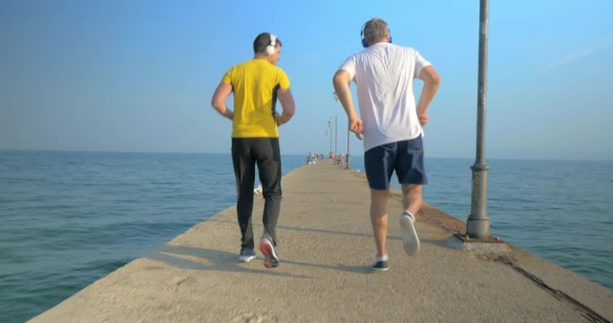 Steadicam shot of senior and young man doing backward exercise on water-front. They run skipping along the pier