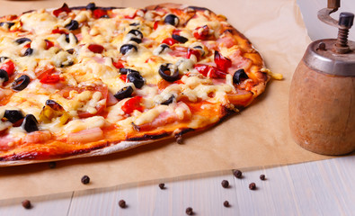 Homemade pizza out of the oven with ham, olives, tomato and cheese on baking paper. On the table in light wood. Pepper mill and spices