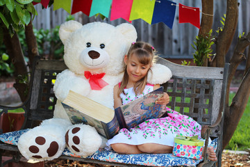 Little girl with her friend huge white bear are reading together a fairy tail book in a garden