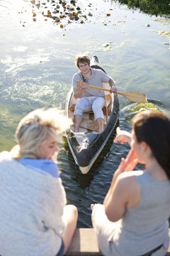 Two female friends sitting on pier pointing at young man rowing
