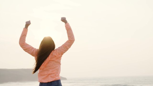 Happy woman raising arms for celebrating workout success and freedom towards the sea on sunset. Successful female athlete shaking arms towards the sun.