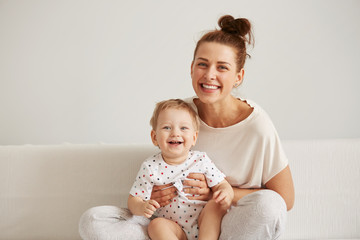 Charming brunette woman playing with her baby while sitting on a bed in her apartment at the...
