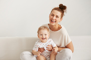 Mother and baby laugh together at home. They are sitting on the sofe in a brightly lit living room at the weekend together, lazy morning, warm and cozy scene. Selective focus