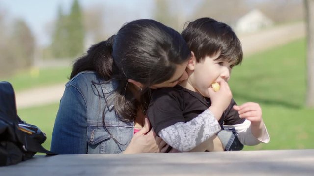 Mother kisses her son as they sit outside at a park bench and he eats sliced apple on a warm spring day.  Recorded hand-held in 4K at 60fps.