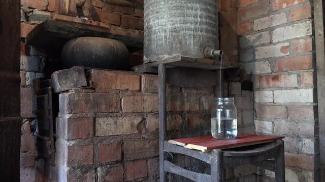 Home made illegal alcohol distillery liquid flow to glass from rusty barrel in secret village room. Static shot. 4K
