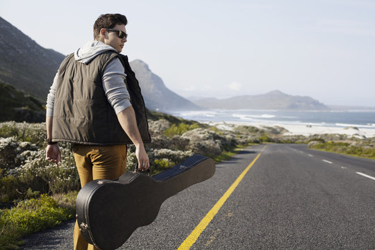 Rear view of young man walking on coastal road carrying guitar case, Cape Town, Western Cape, South Africa - Powered by Adobe