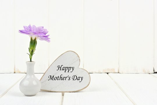 Happy Mother's Day heart tag with small vase and single purple carnation flower against a white wood background