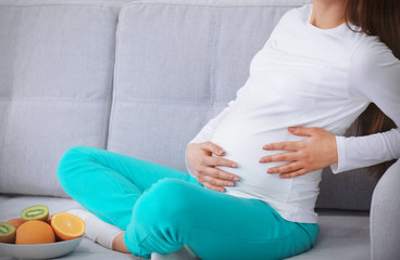 Pregnant woman sitting on sofa with plate of fresh fruits indoors