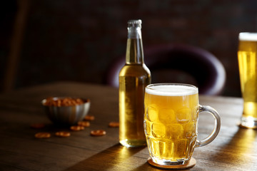 Glass mug of light beer with bottle and snacks on wooden table