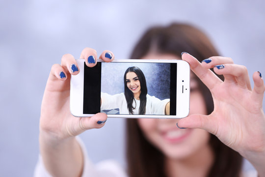 Young attractive girl making photo by her self with mobile phone on grey background, close up