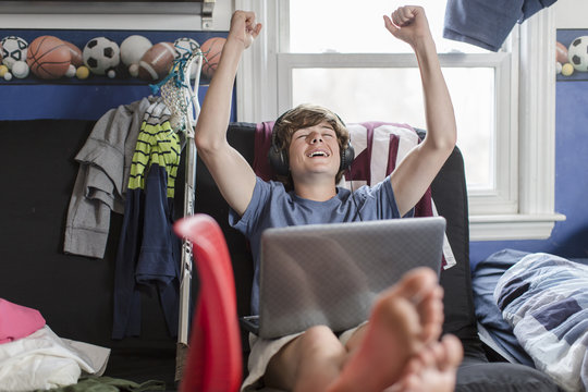 Teenage boy sitting on chair using laptop computer, arms raised