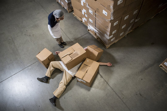 Woman looking at man lying on floor covered by cardboard boxes in warehouse