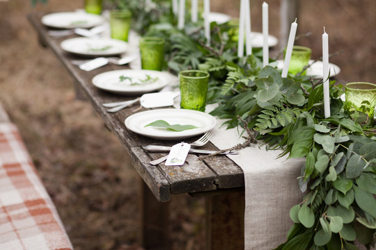 Weddind table setting with white plates and green glasses decorated with white candles, green leaves and eucalyptus and linen tablecloth outdoors