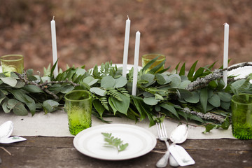 Weddind table setting with white plates and green glasses decorated with white candles, green leaves and eucalyptus outdoors