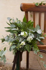 Green bouquet of eucalyptus, ivy, fern, aspidistra, ruskus, date fruit, pink, ranunkulyus with flax box, green and brown velvet ribbons on white stand and brown on wooden chair and beige background