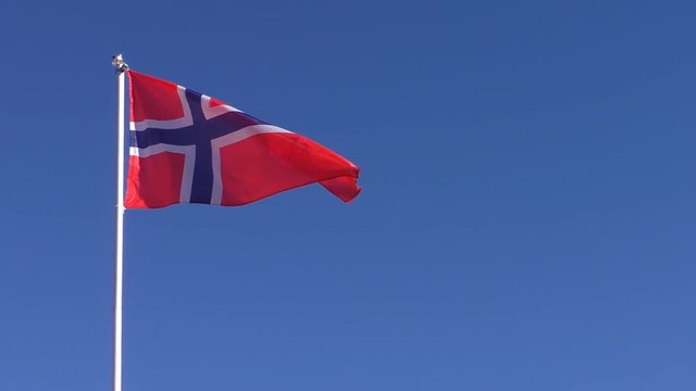 Red with blue cross Norwegian national flag is slowly waving in the blue clear sky on white flagpole