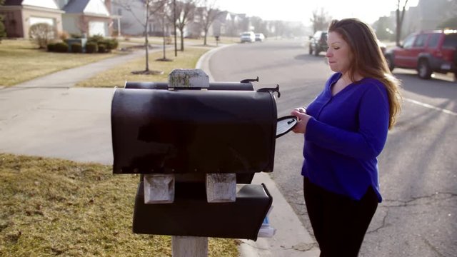 Brunette in blue sweater, collecting letters from her mailbox in a typical suburb of the USA's Mid West.  Early morning sunlight.  Hand held camera, looking down street, recorded in 4K.
