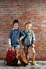 Two stylish little girls with backpacks on wall background